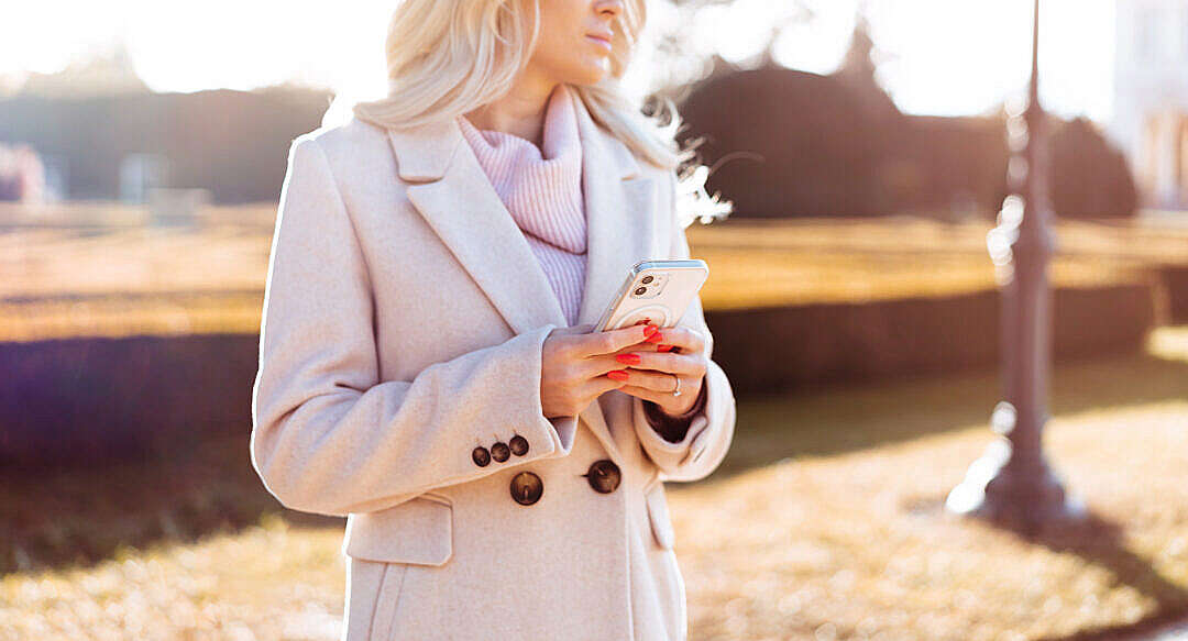 Download Woman in Coat Holding Her White Smartphone in Hands During Sunny Fall FREE Stock Photo