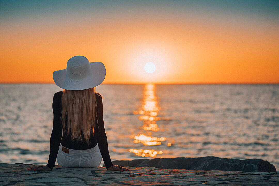 Woman Sitting on a Rocky Shore by the Sea and Watching Sunset